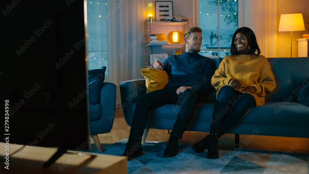 Happy Diverse Young Couple Watching Comedy on TV while Sitting on a Couch, they Laugh and Enjoy Show. Handsome Caucasian Boy and Black Girl in Love Spending Time Together in the Cozy Apartment.