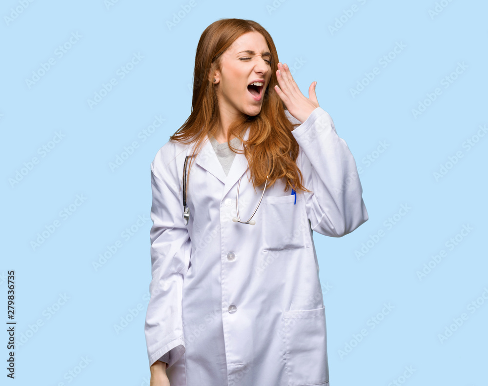 Redhead doctor woman yawning and covering wide open mouth with hand on isolated blue background Redhead doctor woman yawning and covering wide open mouth with hand on isolated blue background