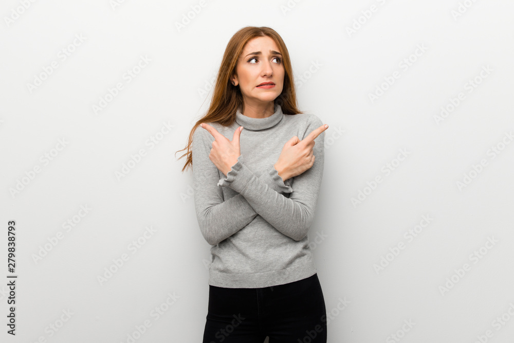Redhead girl over white wall pointing to the laterals having doubts Redhead girl over white wall pointing to the laterals having doubts