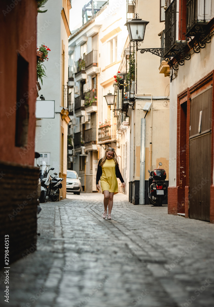 Summer outdoor portrait of young beautiful woman in yellow dress. Cheerful blond girl walking between old stone houses. Enjoying vacation in Portugal. Travel and active lifestyle concept