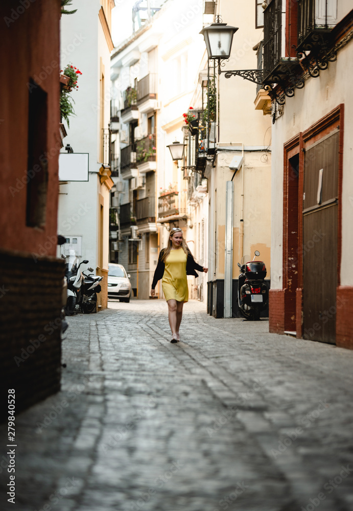 Summer outdoor portrait of young beautiful woman in yellow dress. Cheerful blond girl walking between old stone houses. Enjoying vacation in Portugal. Travel and active lifestyle concept