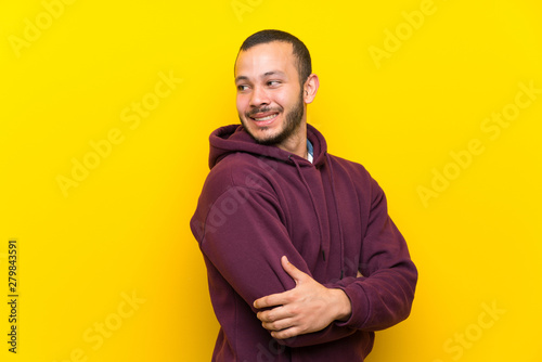 Colombian man with sweatshirt over yellow wall with arms crossed and happy