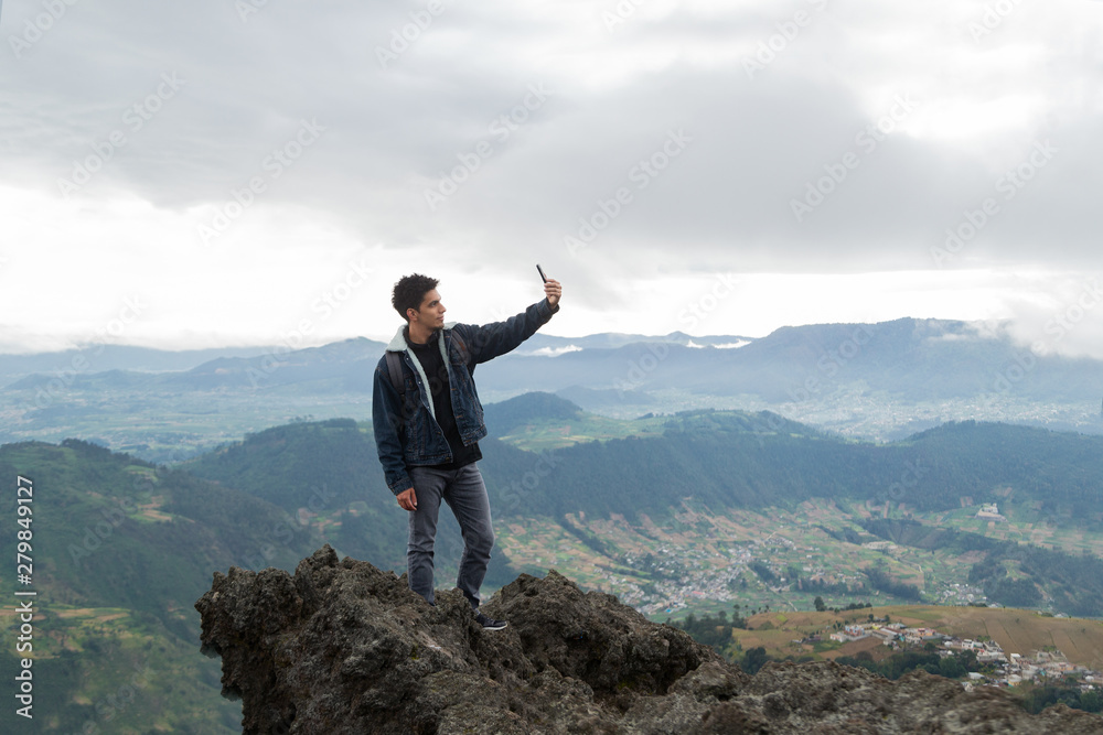 Handsome Hispanic hiker taking a selfie on top of a mountain with the town and mountains behind him- Traveler in Guatemala Handsome Hispanic hiker taking a selfie on top of a mountain with the town and mountains behind him- Traveler in Guatemala
