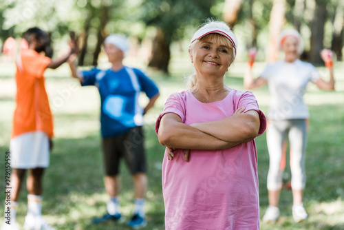 cheerful senior woman standing with crossed arms near multicultural pensioners