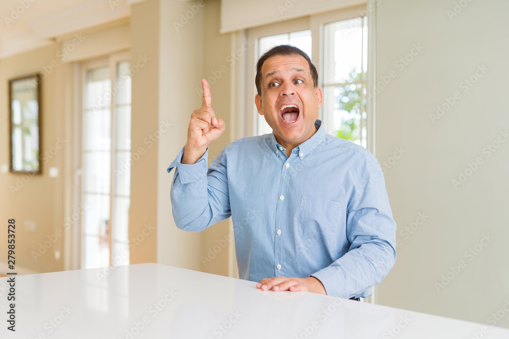 Middle age man sitting at home pointing finger up with successful idea. Exited and happy. Number one. Middle age man sitting at home pointing finger up with successful idea. Exited and happy. Number one.