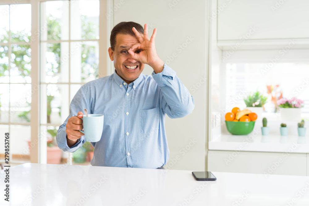 Middle age business man drinking coffee in the morning at home with happy face smiling doing ok sign with hand on eye looking through fingers