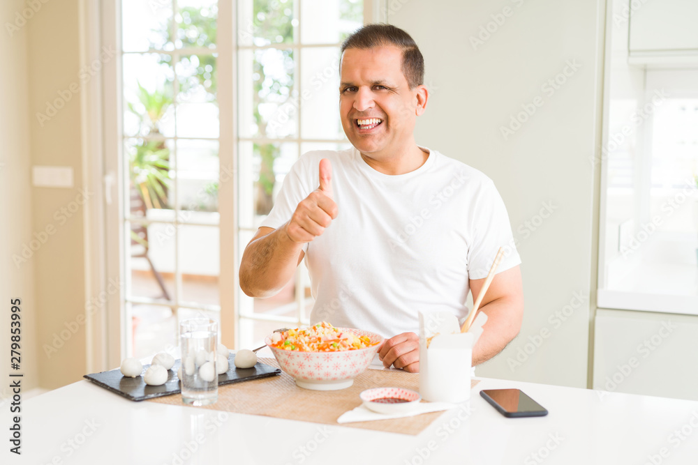 Middle age man eating asian food with chopsticks at home doing happy thumbs up gesture with hand. Approving expression looking at the camera with showing success. Middle age man eating asian food with chopsticks at home doing happy thumbs up gesture with hand. Approving expression looking at the camera with showing success.