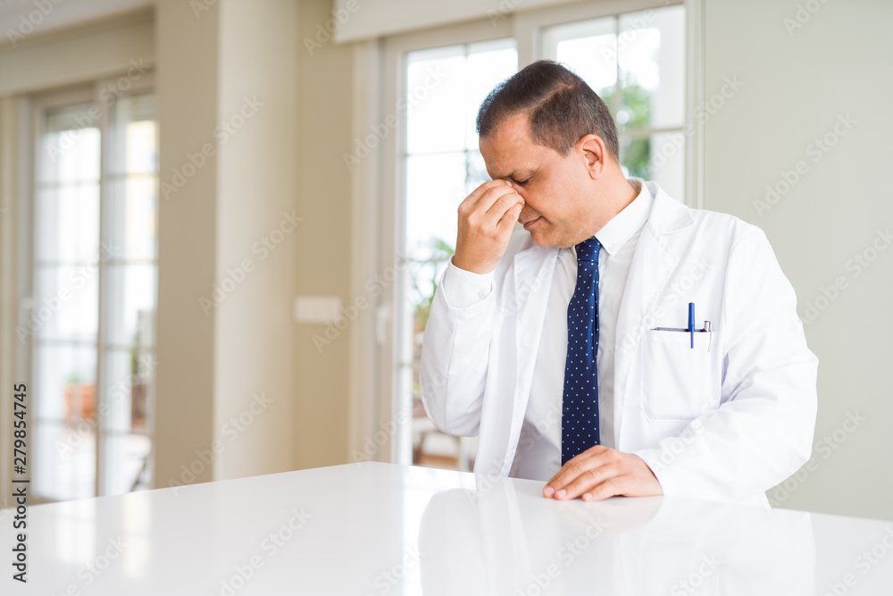 Middle age doctor man wearing medical coat at the clinic tired rubbing nose and eyes feeling fatigue and headache. Stress and frustration concept.