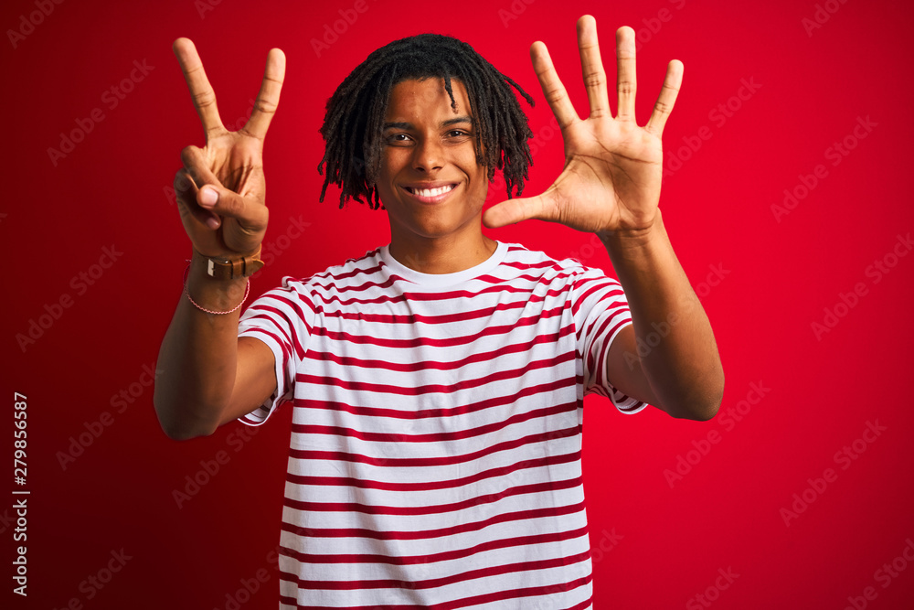Young afro man with dreadlocks wearing striped t-shirt standing over isolated red background showing and pointing up with fingers number seven while smiling confident and happy. Young afro man with dreadlocks wearing striped t-shirt standing over isolated red background showing and pointing up with fingers number seven while smiling confident and happy.