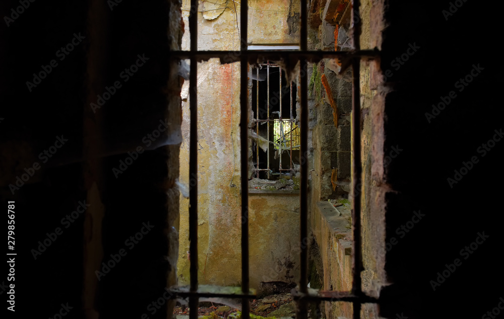 A photo of a dirty run down prison cell ruins. The bars are rusted badly and the walls are badly decayed.