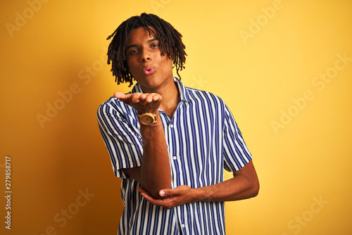 Afro man with dreadlocks wearing casual striped t-shirt over isolated yellow background looking at the camera blowing a kiss with hand on air being lovely and sexy. Love expression. © Krakenimages.com