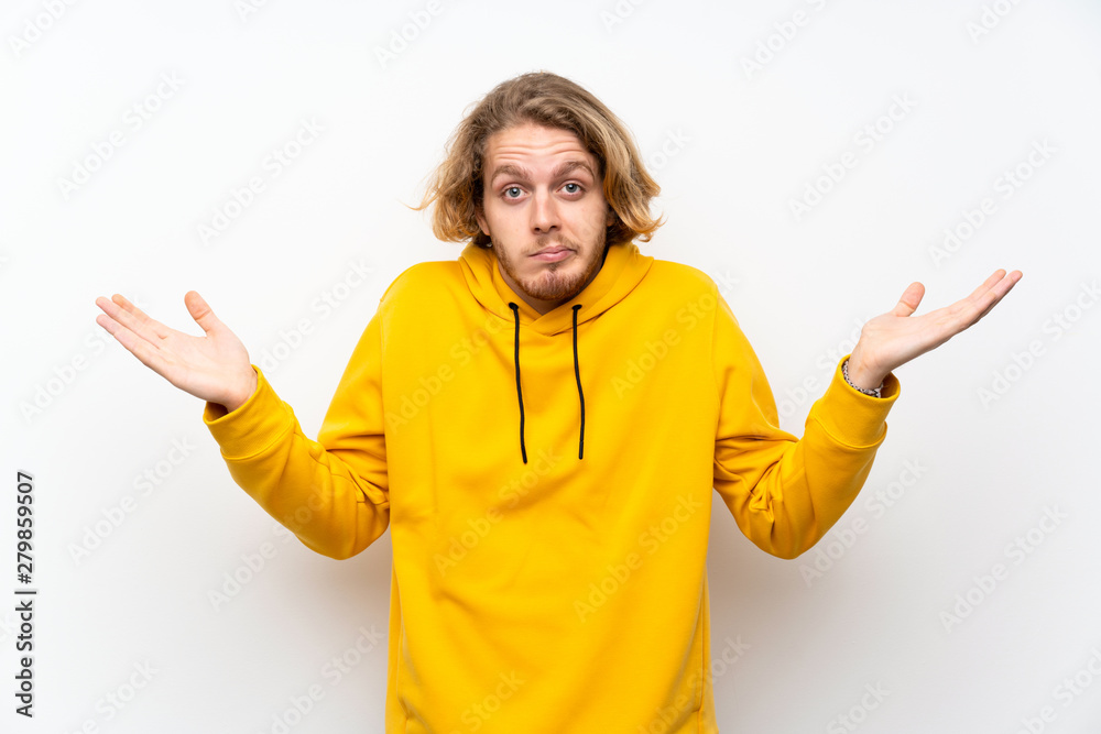 Blonde man with sweatshirt over white wall having doubts while raising hands Blonde man with sweatshirt over white wall having doubts while raising hands