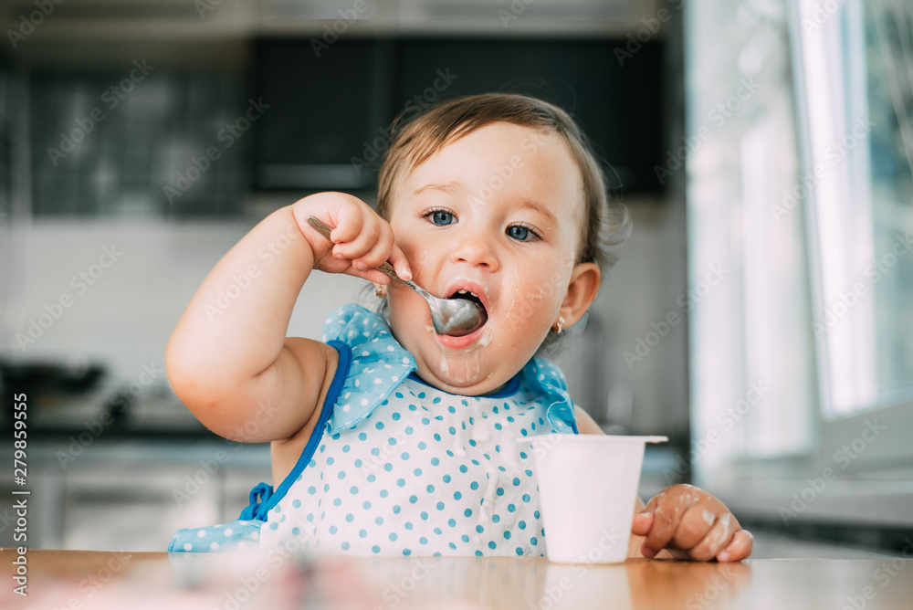 Cute, funny girl eating yogurt during the day in the kitchen in a blue dress Cute, funny girl eating yogurt during the day in the kitchen in a blue dress