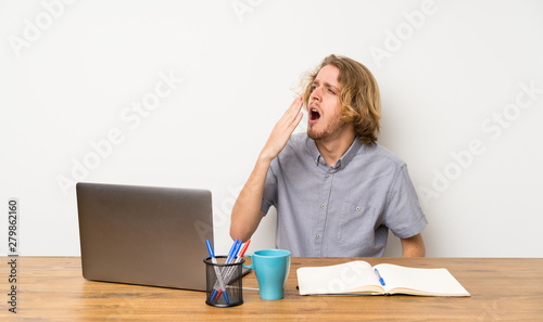 Blonde man with a laptop yawning and covering wide open mouth with hand