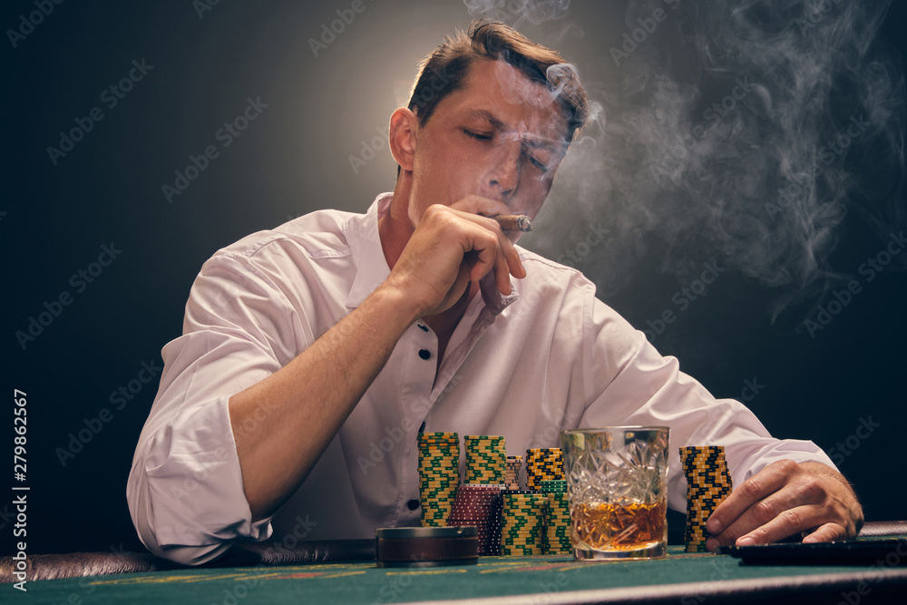 Handsome emotional man is playing poker sitting at the table in casino against a white spotlight. Handsome emotional man is playing poker sitting at the table in casino against a white spotlight.
