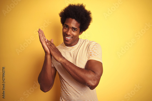 American man with afro hair wearing striped t-shirt standing over isolated yellow background clapping and applauding happy and joyful, smiling proud hands together