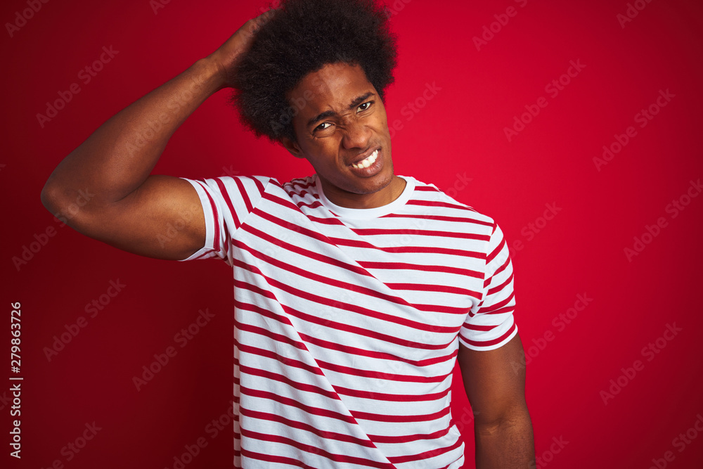 Young african american man with afro hair wearing striped t-shirt over isolated red background confuse and wonder about question. Uncertain with doubt, thinking with hand on head. Pensive concept. Young african american man with afro hair wearing striped t-shirt over isolated red background confuse and wonder about question. Uncertain with doubt, thinking with hand on head. Pensive concept.