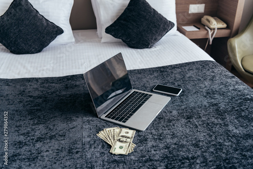laptop with blank screen, smartphone and dollar banknotes on bed in hotel room