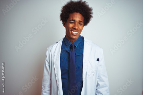 Young african american doctor man wearing coat standing over isolated white background winking looking at the camera with sexy expression, cheerful and happy face.