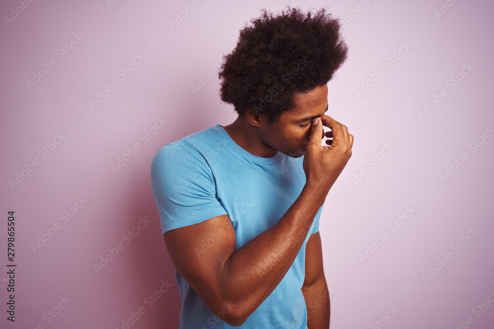 African american man with afro hair wearing blue t-shirt standing over isolated pink background tired rubbing nose and eyes feeling fatigue and headache. Stress and frustration concept. African american man with afro hair wearing blue t-shirt standing over isolated pink background tired rubbing nose and eyes feeling fatigue and headache. Stress and frustration concept.