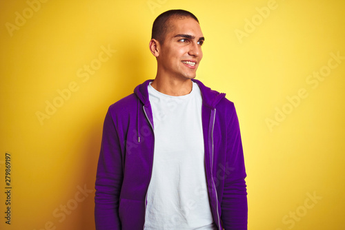 Young handsome man wearing purple sweatshirt over yellow isolated background looking away to side with smile on face, natural expression. Laughing confident.