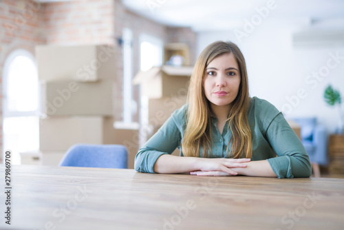 Young woman sitting on the table with cardboard boxes behind her moving to new home with serious expression on face. Simple and natural looking at the camera.