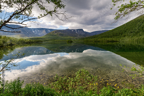 Beautiful view of a mountain lake Small Vudyavr with reflection of clouds in the water. In the foreground are flowers. Khibiny, Murmansk region, Kola Peninsula, Russia. photo