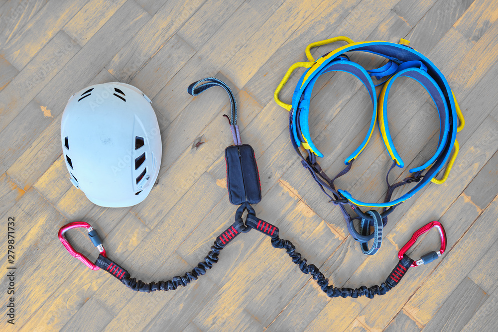 Flat lay of via ferrata gear (white helmet, two lanyards with shock absorber system, climbing harness) on wooden background with light orange spots.