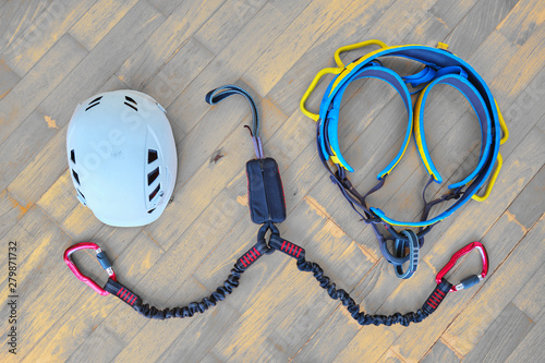 Flat lay of via ferrata gear (white helmet, two lanyards with shock absorber system, climbing harness) on wooden background with light orange spots.