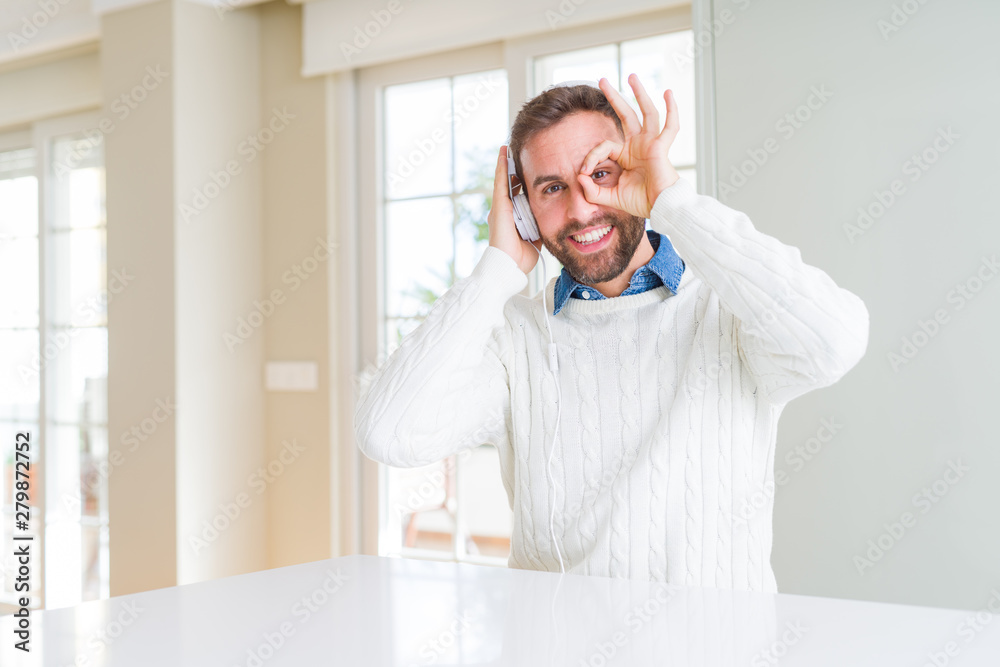 Handsome man wearing headphones and listening to music with happy face smiling doing ok sign with hand on eye looking through fingers