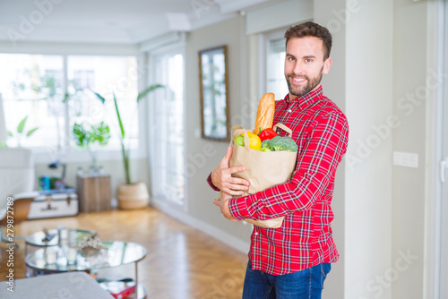 Handsome man holding paper bag full of fresh groceries at home photo