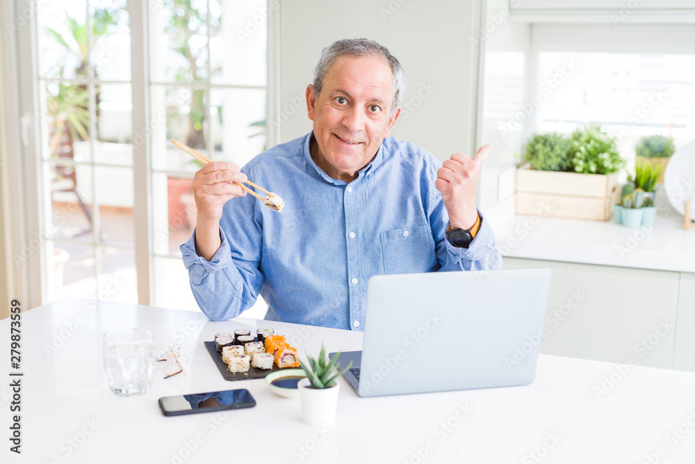 Handsome business senior man eating delivery sushi while working using laptop pointing and showing with thumb up to the side with happy face smiling