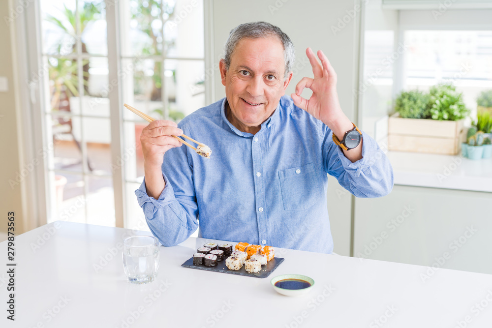Handsome senior man eating take away sushi using chopsticks at home doing ok sign with fingers, excellent symbol Handsome senior man eating take away sushi using chopsticks at home doing ok sign with fingers, excellent symbol