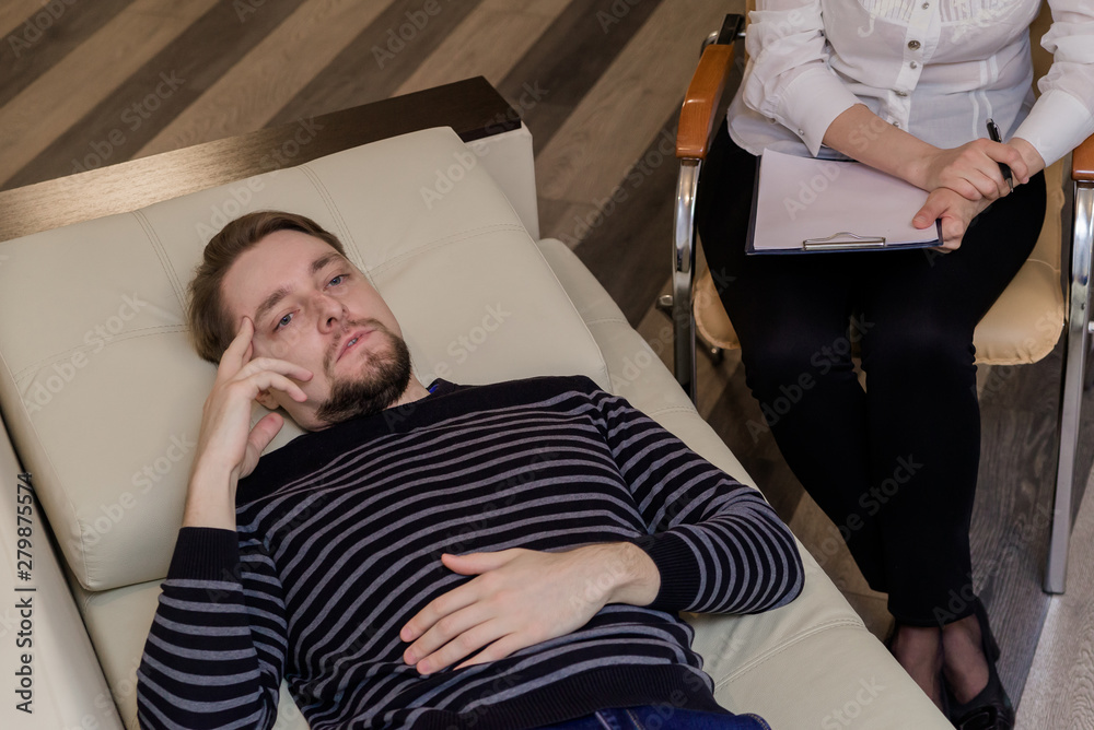 Fototapeta premium Young man at the psychotherapist, lying on couch, emotionally speaking, discussing with psychotherapist his problems, practitioner making some notes