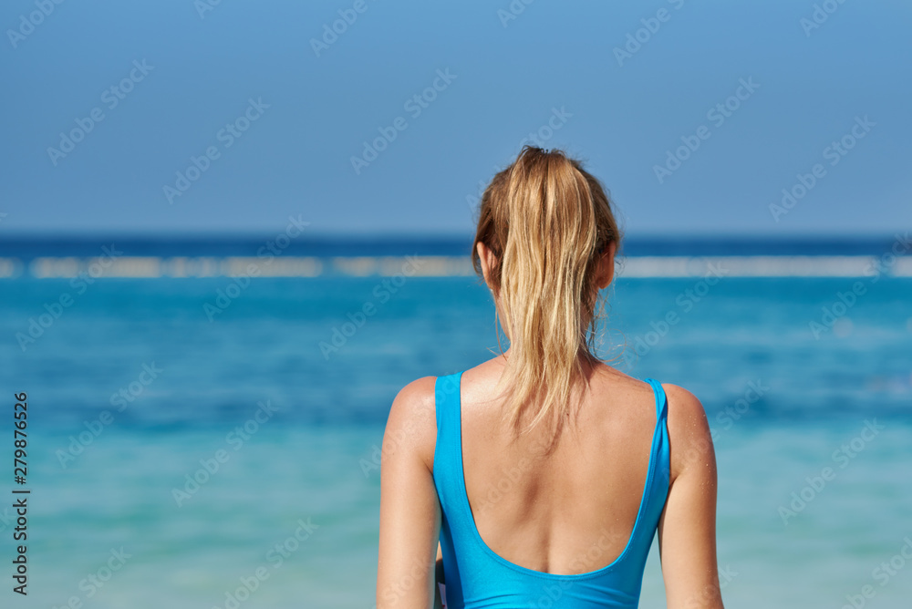 Blonde girl in swimsuit against blue sea water on beach during summer vacation. Back view.