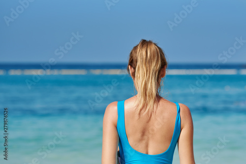 Blonde girl in swimsuit against blue sea water on beach during summer vacation. Back view. © Artem
