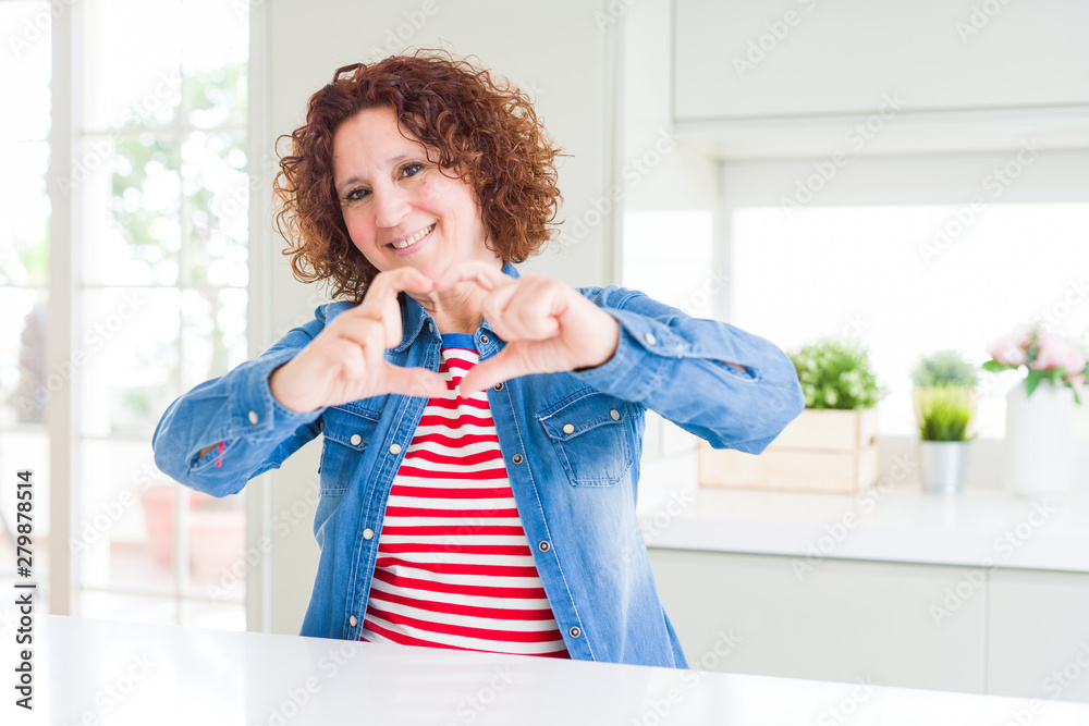 Middle age senior woman with curly hair wearing denim jacket at home smiling in love doing heart symbol shape with hands. Romantic concept.