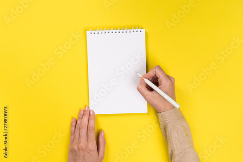 Woman's hands with perfect manicure holding pencil and  notepad as mockup for your design. Yellow background, flat lay style.