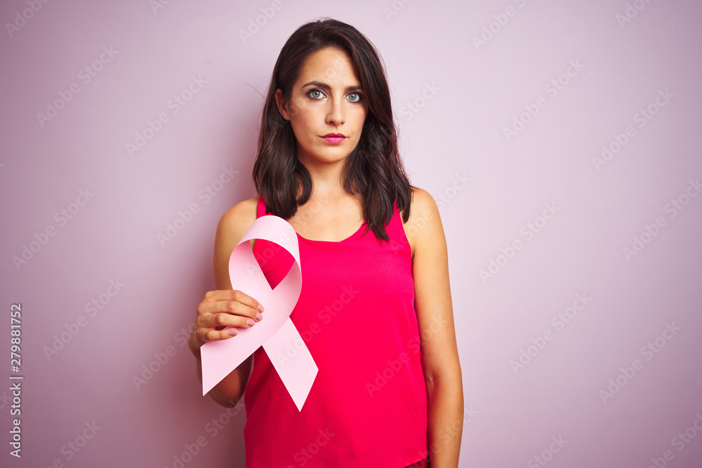 Young beautiful woman holding cancer ribbon over pink isolated background with a confident expression on smart face thinking serious Young beautiful woman holding cancer ribbon over pink isolated background with a confident expression on smart face thinking serious