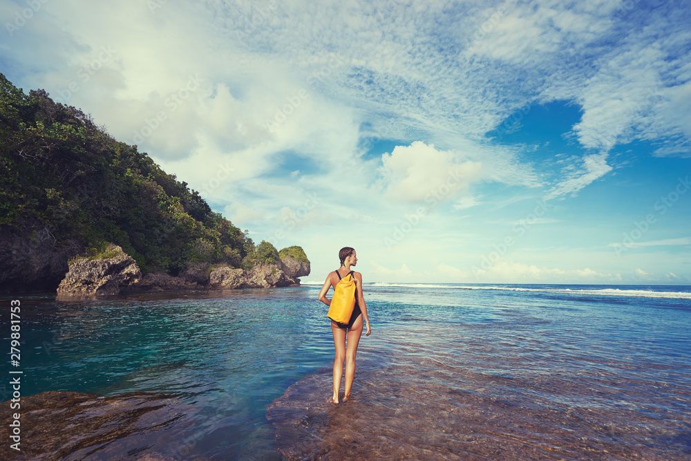 Tropical traveling. Young woman with waterproof backpack walking by low tide sea beach.