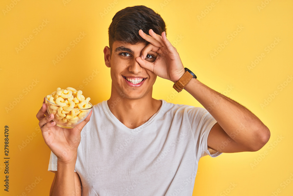 Young indian man holding bowl with extruded corn standing over isolated yellow background with happy face smiling doing ok sign with hand on eye looking through fingers Young indian man holding bowl with extruded corn standing over isolated yellow background with happy face smiling doing ok sign with hand on eye looking through fingers