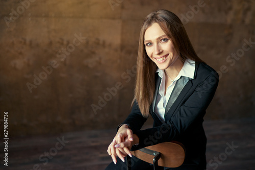 Studio portrait of young beautiful sensual woman in blak suit sitting on wooden chair against dark background.