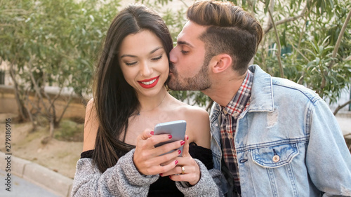 Foto man kissing young woman but she is distracted with phone addiction