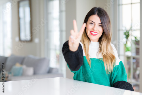 Young beautiful woman wearing winter sweater at home smiling looking to the camera showing fingers doing victory sign. Number two. Free Download Young beautiful woman wearing winter sweater at home smiling looking to the camera showing fingers doing victory sign. Number two. - GFXLoad