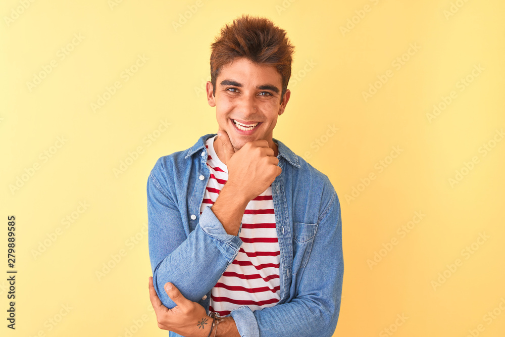 Young handsome man wearing striped t-shirt and denim shirt over isolated yellow background looking confident at the camera smiling with crossed arms and hand raised on chin. Thinking positive. Young handsome man wearing striped t-shirt and denim shirt over isolated yellow background looking confident at the camera smiling with crossed arms and hand raised on chin. Thinking positive.