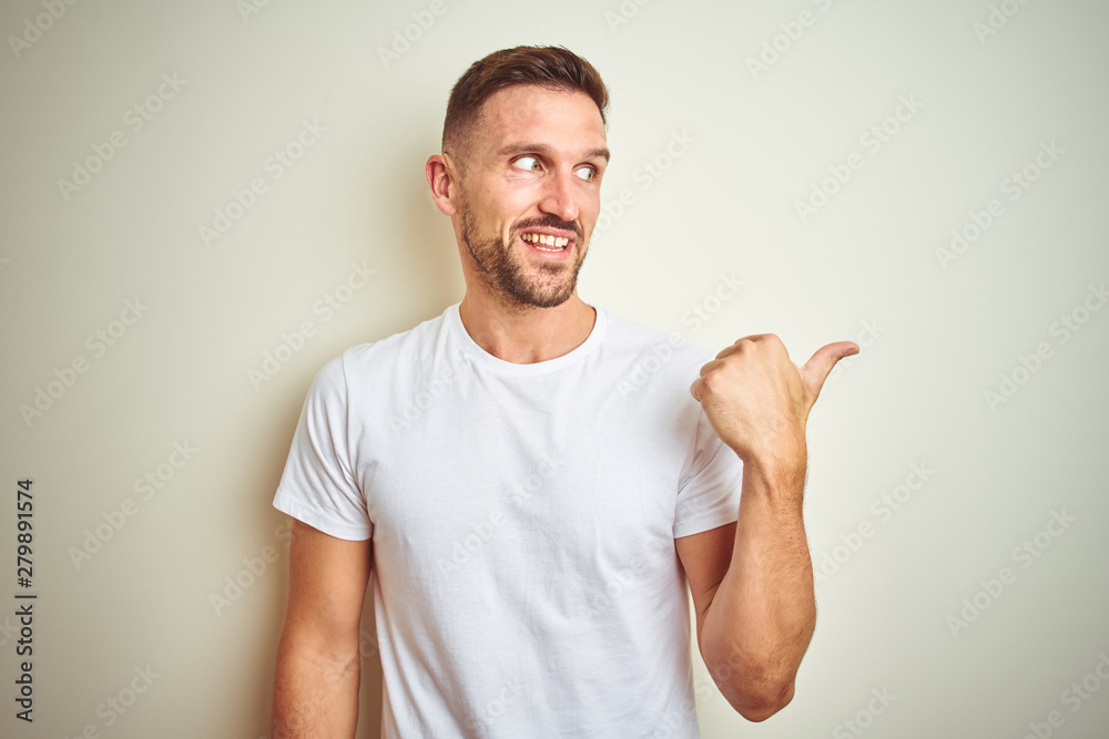 Young handsome man wearing casual white t-shirt over isolated background smiling with happy face looking and pointing to the side with thumb up. Young handsome man wearing casual white t-shirt over isolated background smiling with happy face looking and pointing to the side with thumb up.