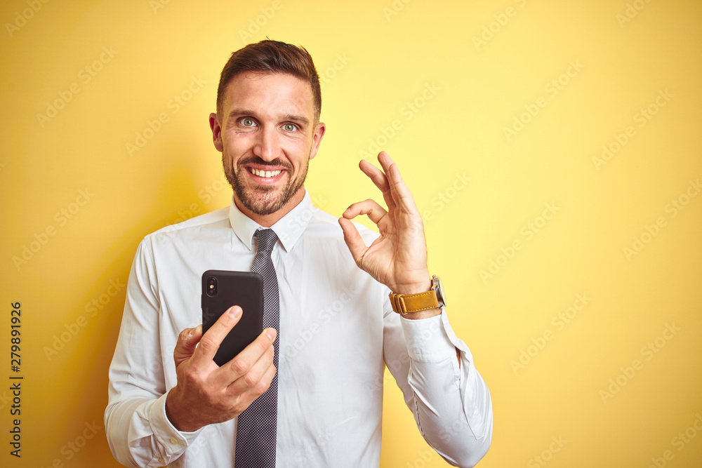 Young handsome business man using smartphone over yellow isolated background doing ok sign with fingers, excellent symbol Young handsome business man using smartphone over yellow isolated background doing ok sign with fingers, excellent symbol