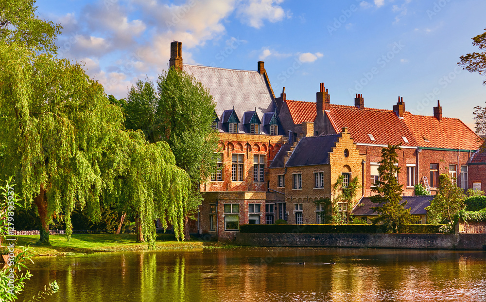 Vintage building over lake of love in Minnewater park in Bruges Belgium near Beguinage monastery of Beguines. Picturesque landscape with green trees sunset time.