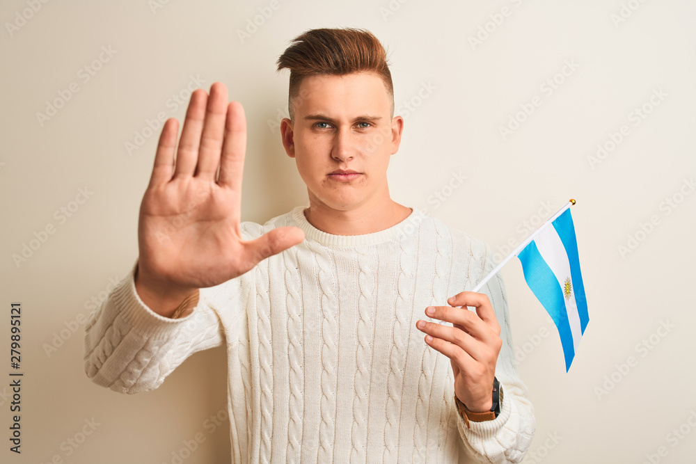 Young handsome man holding Argentinian Argentina flag over isolated white background with open hand doing stop sign with serious and confident expression, defense gesture Young handsome man holding Argentinian Argentina flag over isolated white background with open hand doing stop sign with serious and confident expression, defense gesture