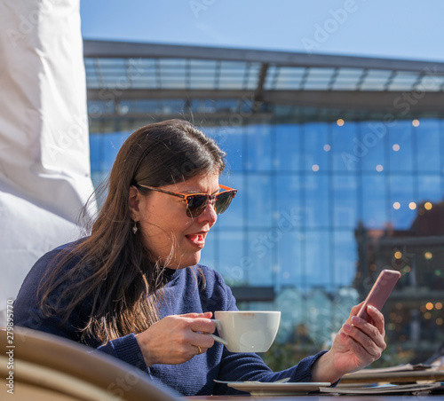 Lady sits at a city cafe outdoors and checks her phone photo